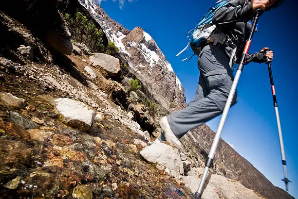 Climber using trekking poles to cross a rocky stream on Mount Kilimanjaro with snow-covered peaks in the background.